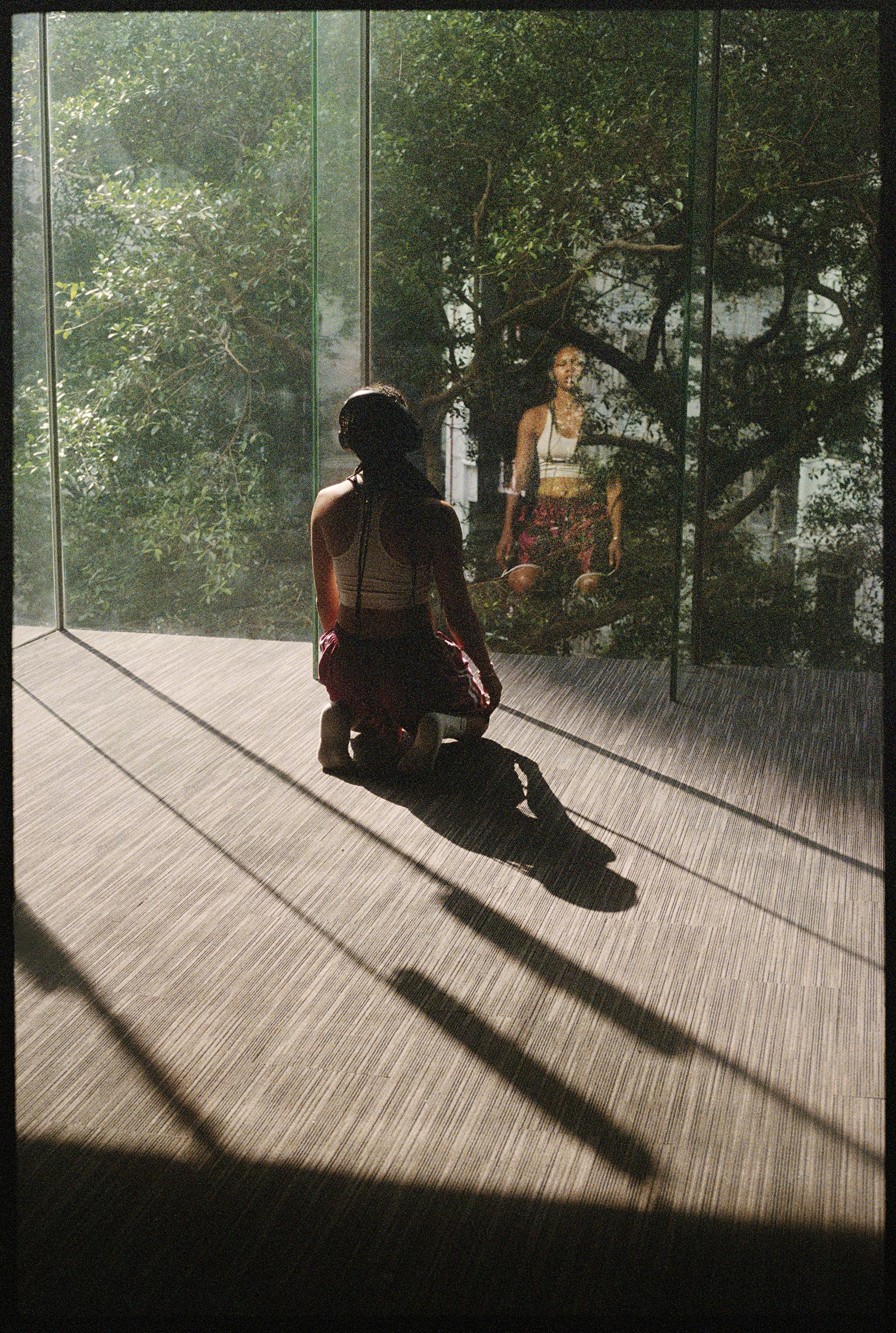 Dancer seated on her knees in the studio, facing the greenery outside the bay windows.