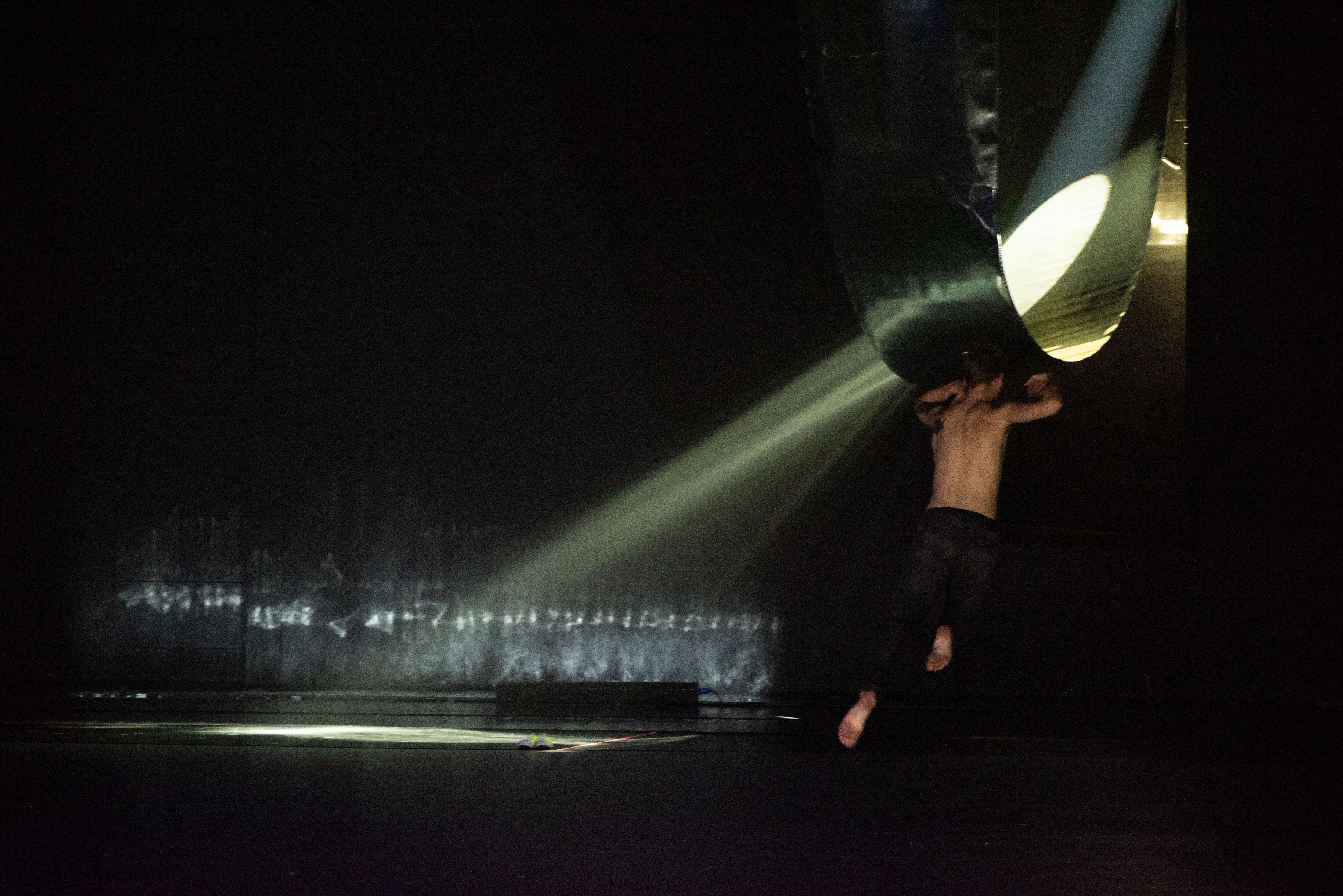 A dancer jumping in front of a structure diffusing a ray of light on a dark stage