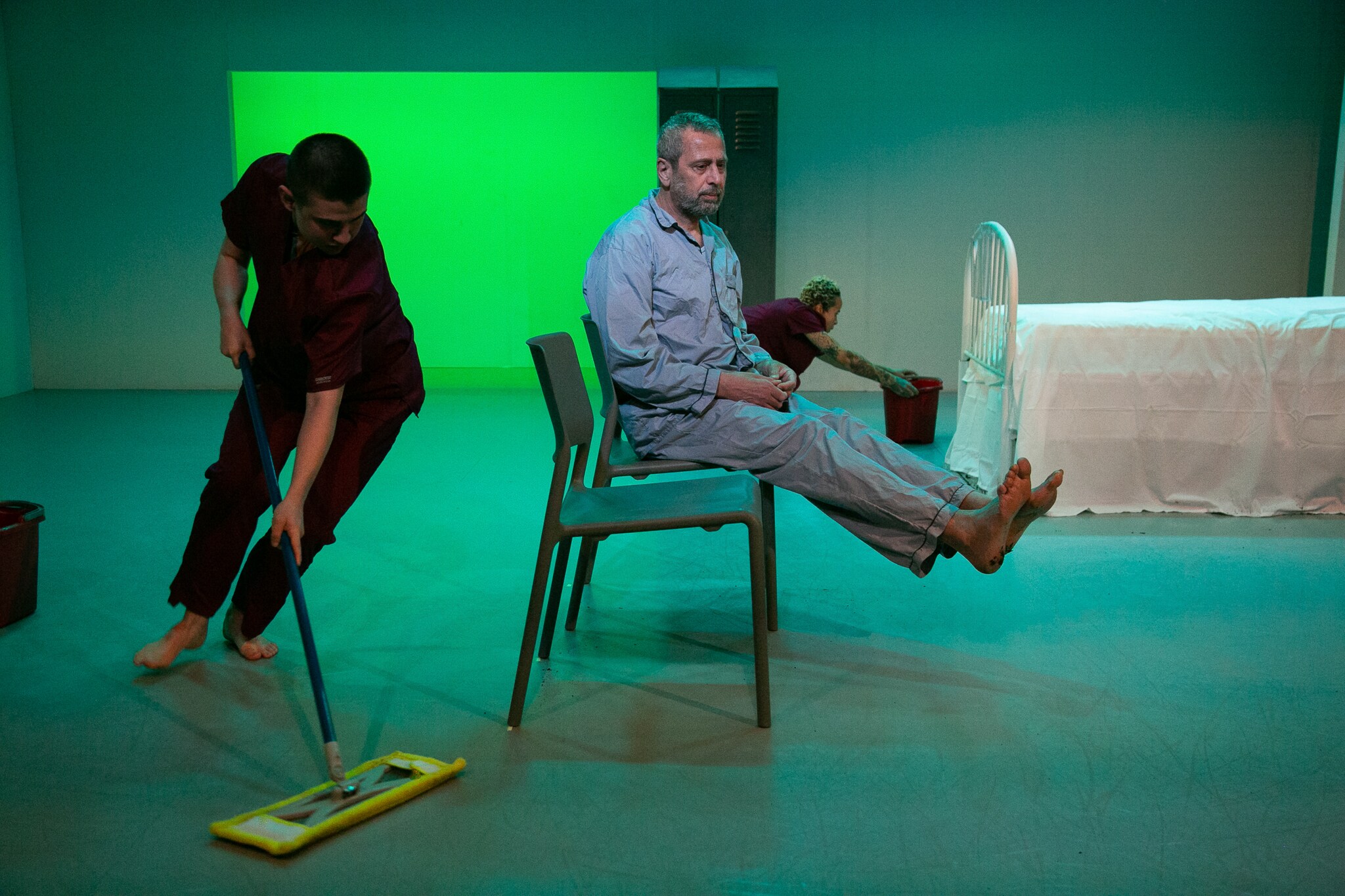 un homme assis sur une chaise dans un hôpital, les pieds en l’air tandis qu’un soignant nettoie le sol