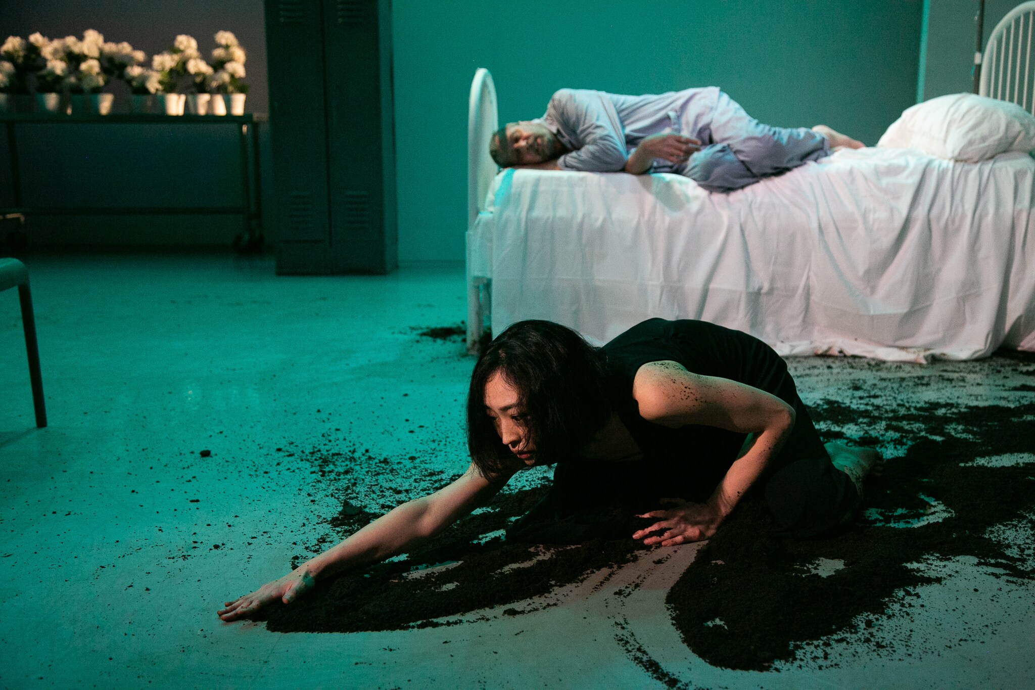 un homme allongé sur un lit d’hôpital avec une femme en robe noire, étalant de la terre sur le sol