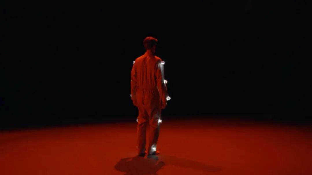Homme de dos, portant des lumières sur son costume, sur une scène baignée de lumière rouge