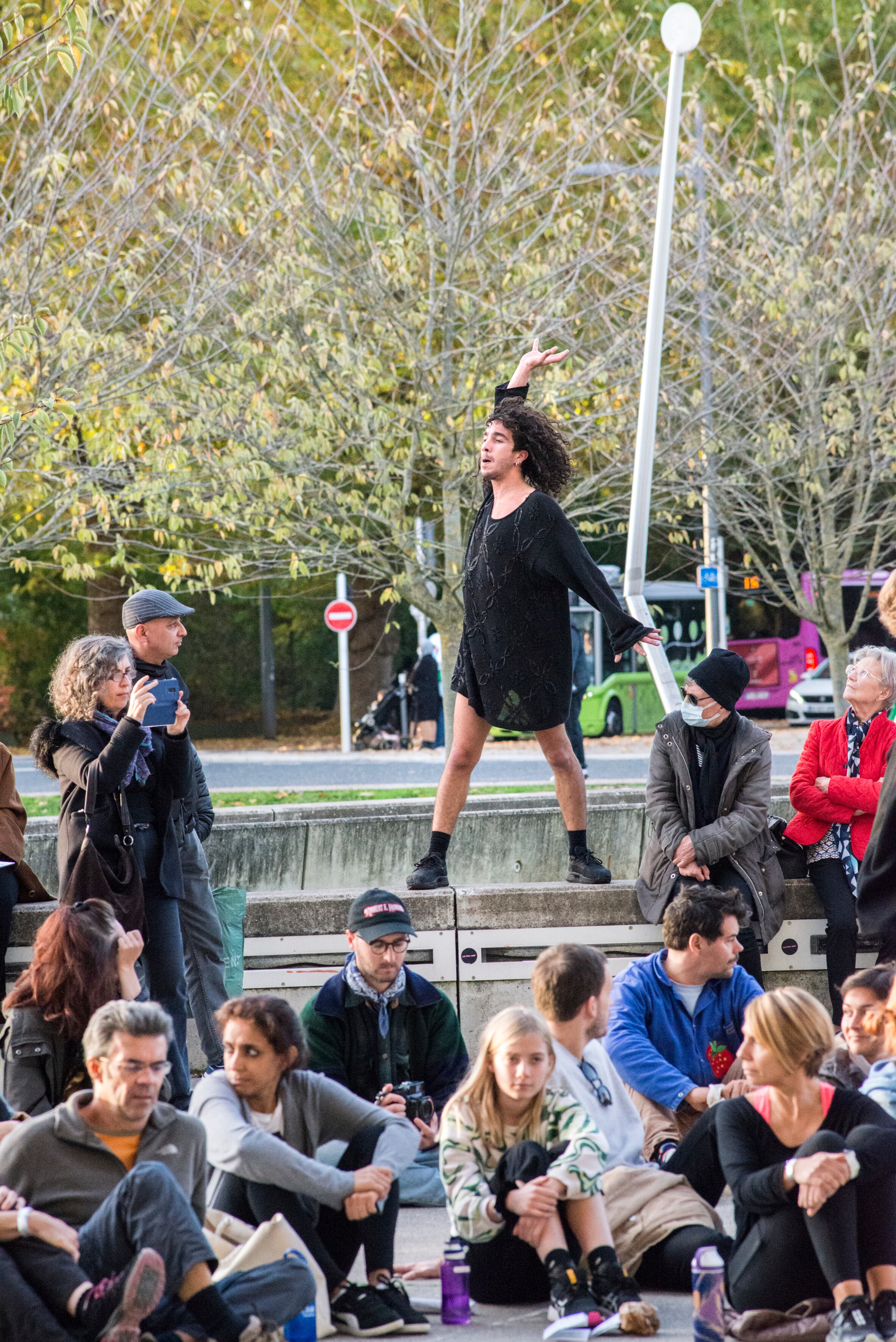 Dancer dressed in a black tunic standing on a concrete wall in the middle of the audience, who are sitting cross-legged