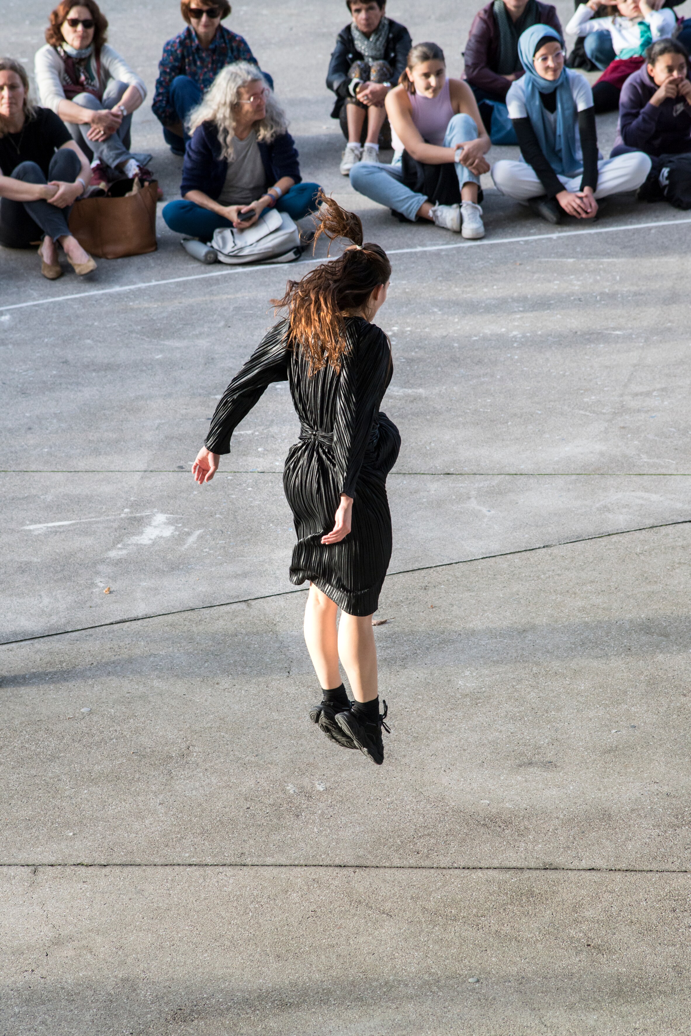 Dancer wearing a black dress, jumping on the forecourt of the Center Pompidou-Metz, facing the audience, who are sitting cross-legged