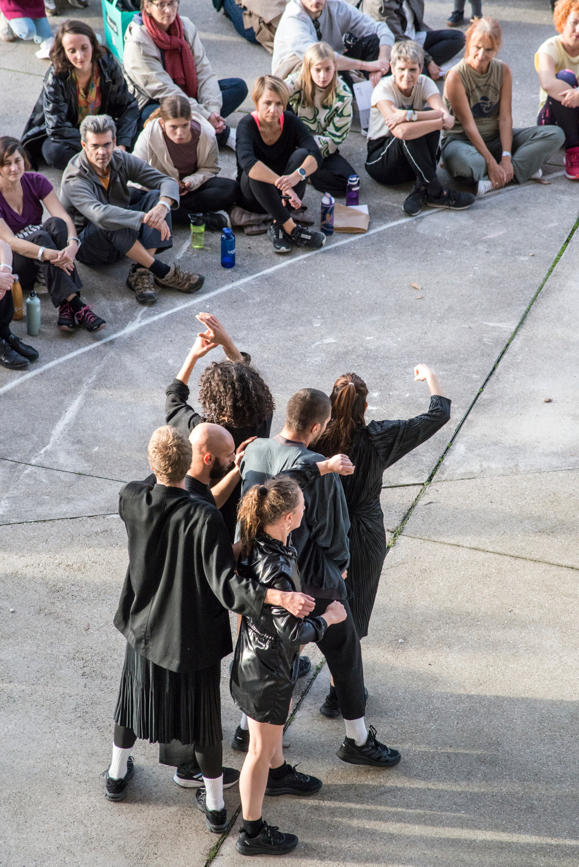 Dancers dressed in black on the forecourt of the Center Pompidou-Metz, facing the audience, who are sitting cross-legged