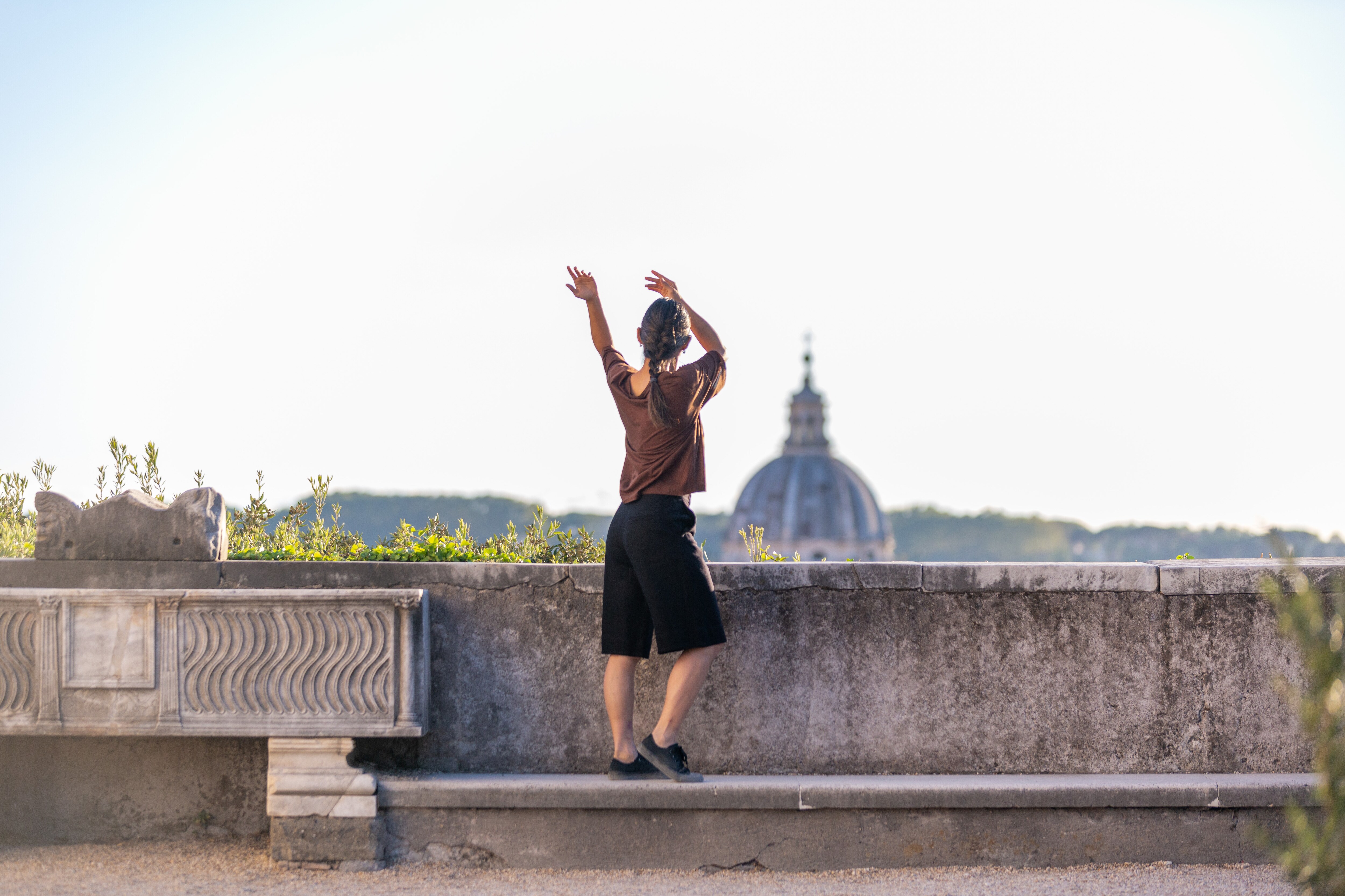 Dancer turned away with arms raised, standing on a low concrete wall