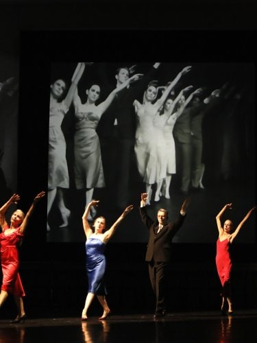 Four women and one man standing still in the foreground. A screen behind them projects dancers in the same position.