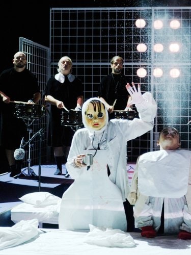 two figures in white costumes, one masked and the other resembling a puppet, in front of musicians