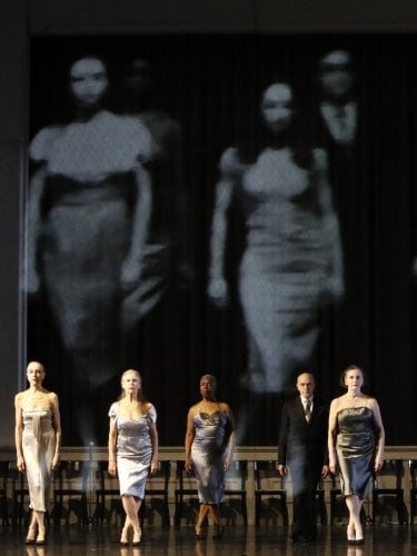 Dancers in elegant costumes on stage, with a projection of blurred black and white silhouettes in the background.