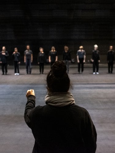 Nacera Belaza de dos et face à elle 12 danseurs en ligne vêtus de noir