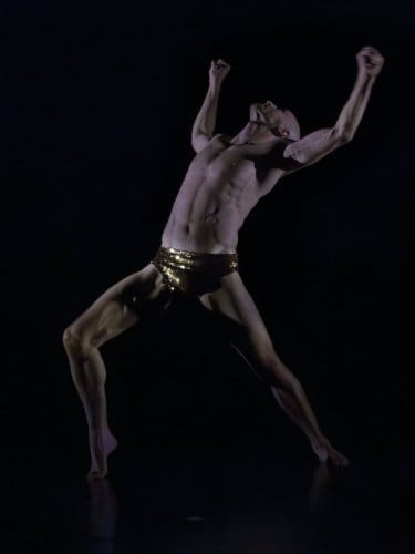 dancer arms raised, on a dark stage