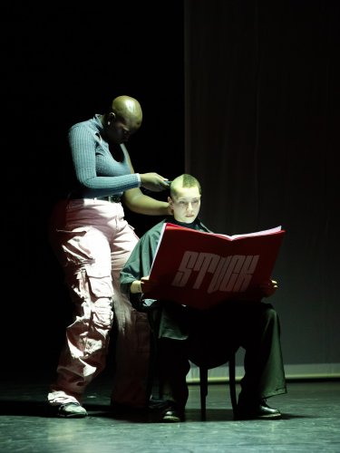 one dancer shaves the head of another who is seated and reading a large red book with "STUCK" written on it