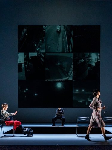 cinq danseurs sur scène, une rangée de chaises pliantes noires et un grand écran affichant une grille de flux vidéo de surveillance sombres