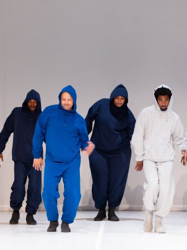 Cinq danseurs en survêtements bleu foncé, bleu vif, et gris clair, dansant en groupe, à droite de l'image un décor peint d'un paysage enneigé et rocheux