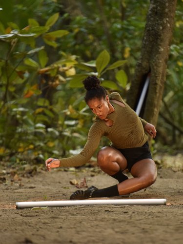danseuse accroupie sur un sol terreux dans une forêt luxuriante, le bras tendu vers un tube blanc posé au sol