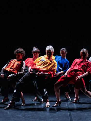 Groupe de danseuses en vêtements colorés et talons hauts dans une pose dynamique vers l'arrière, sur une scène sombre