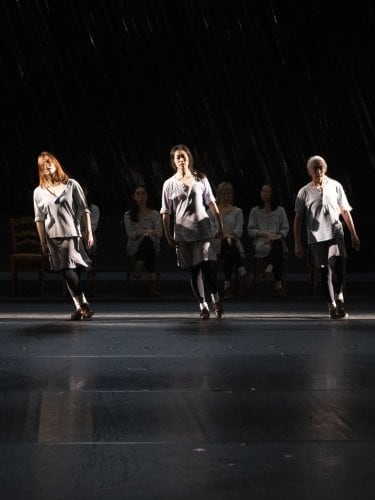 Dancers on a dark stage, some standing in the light, others seated in the shadow, wearing light tunics