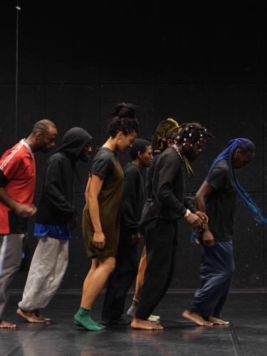 Six dancers, mostly barefoot, moving in a line across a dark stage