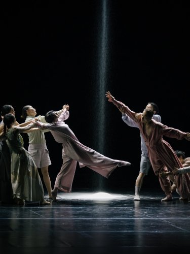 Danseurs en costumes fluides sur une scène sombre, avec un faisceau de lumière et du sable tombant au centre