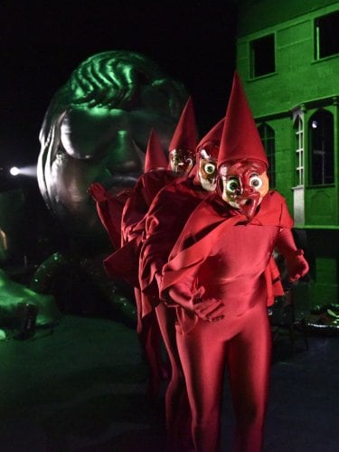 Personnages masqués en costumes rouges vifs et chapeaux pointus, marchant en ligne devant un bâtiment éclairé en vert