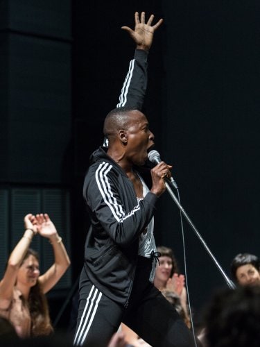 A performer in a black tracksuit sings passionately into a microphone, one arm raised, with an audience visible in the background, some with their hands raised