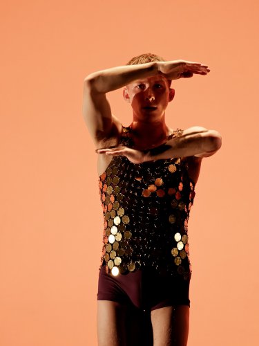 Dancer whose arms are bent in front of his face, background orange. 