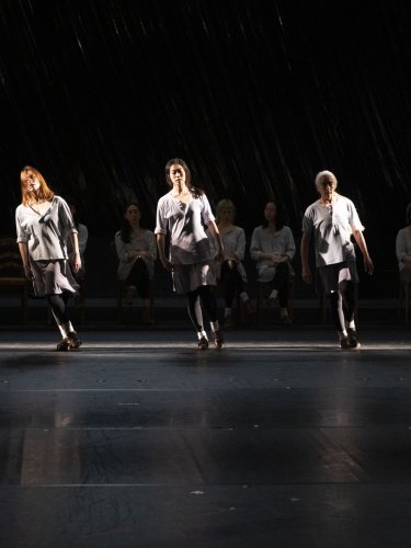 Dancers on a dark stage, some standing in the light, others seated in the shadow, wearing light tunics
