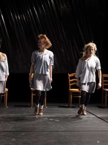 Dancers on a dark stage, some standing in the light, others seated in the shadow, wearing light tunics