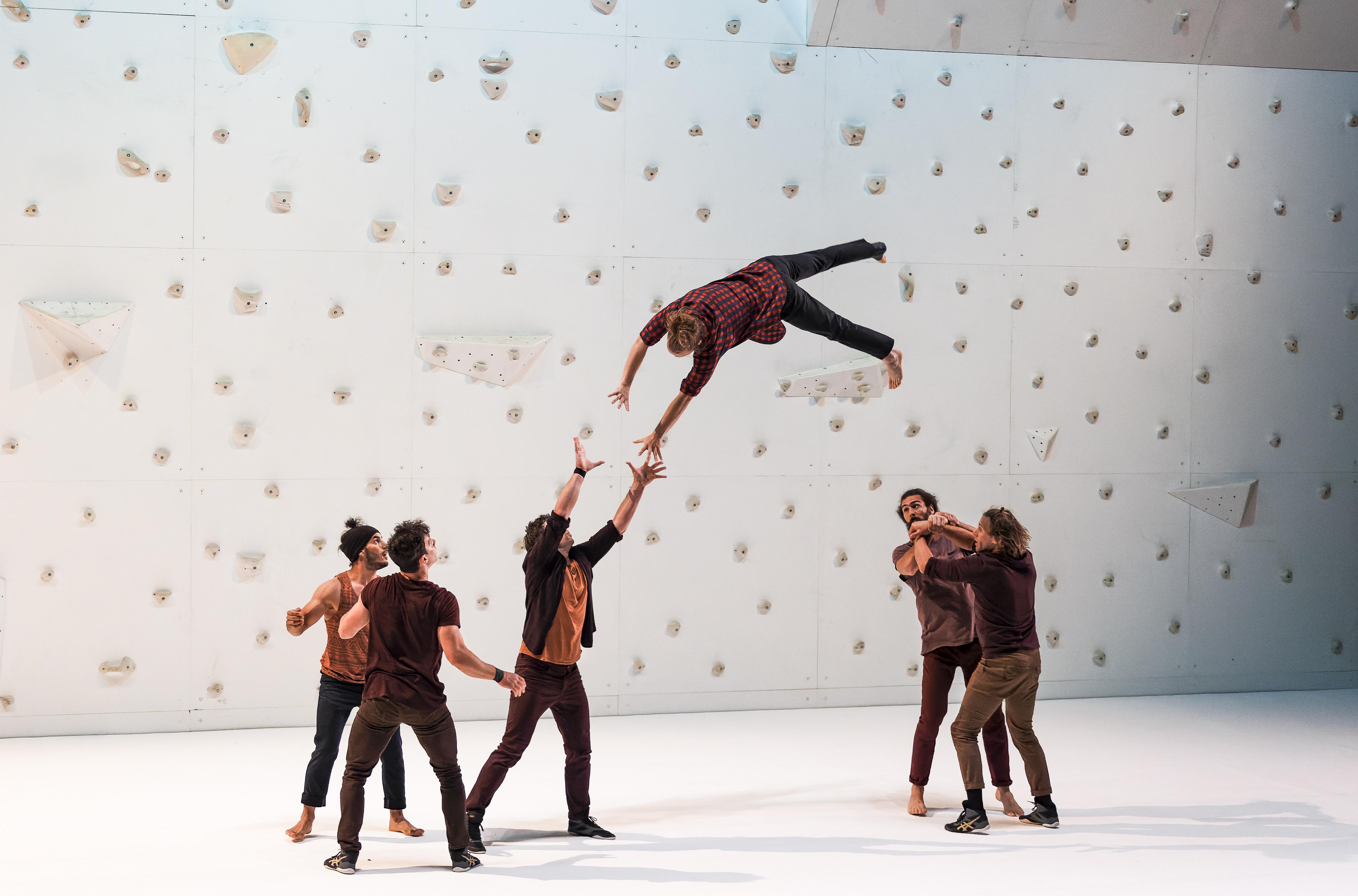 Dancers in front of a white climbing wall throwing a dancer in the air 