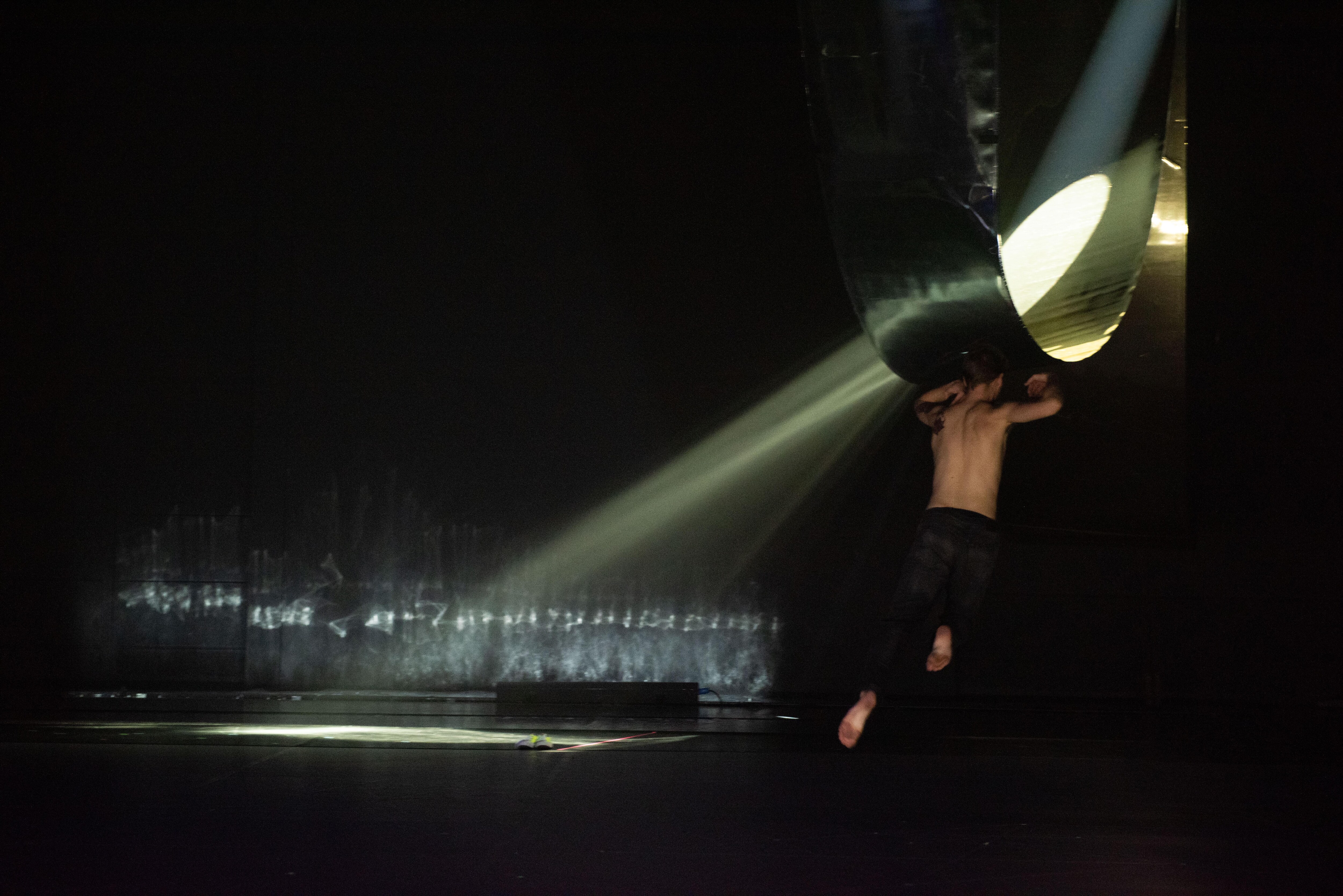 在黑暗的舞台上，一位舞者在滲透出一絲光線的建築物前跳躍