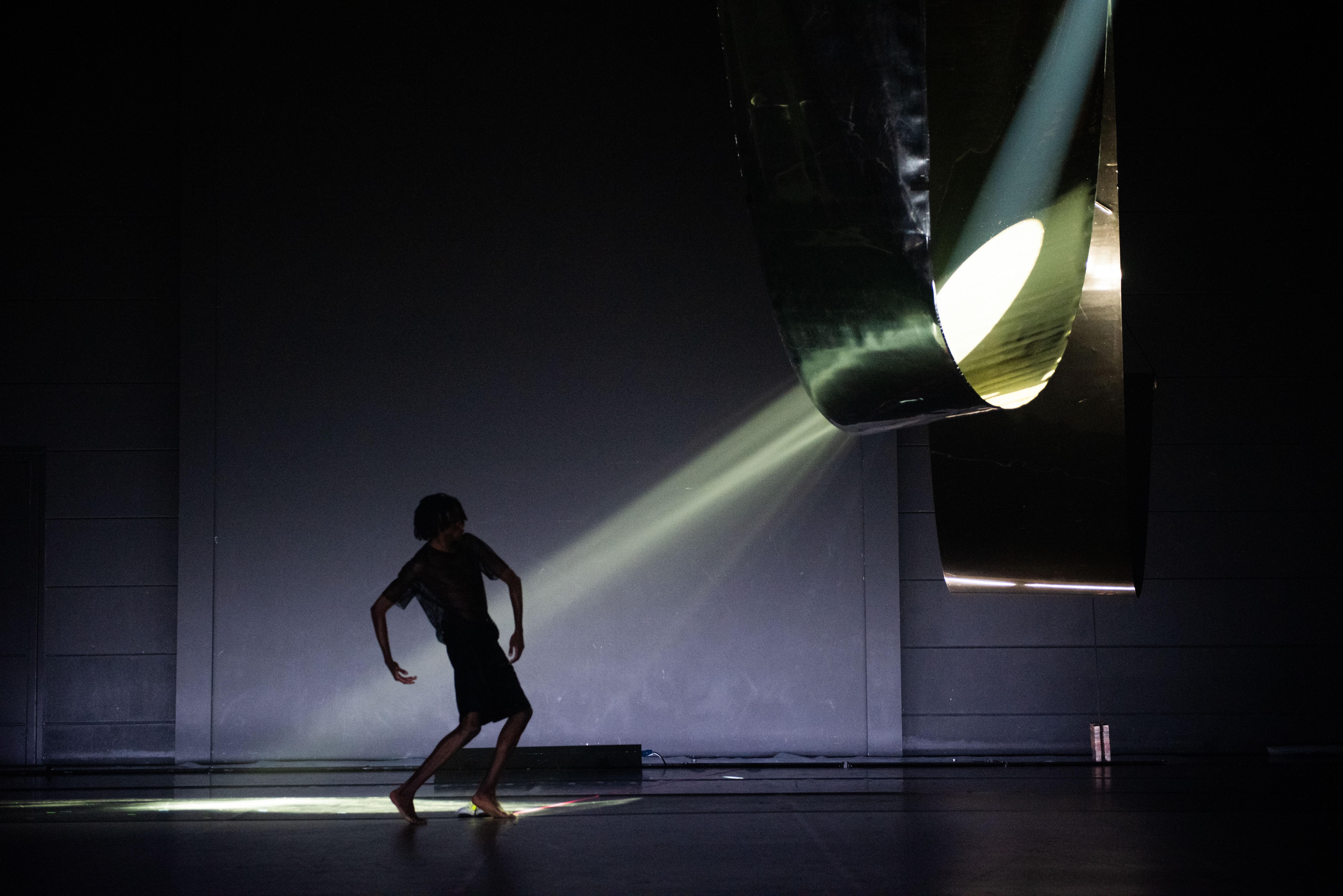 「一位舞者在黑暗的舞台上，面對著一個散發光線的結構」