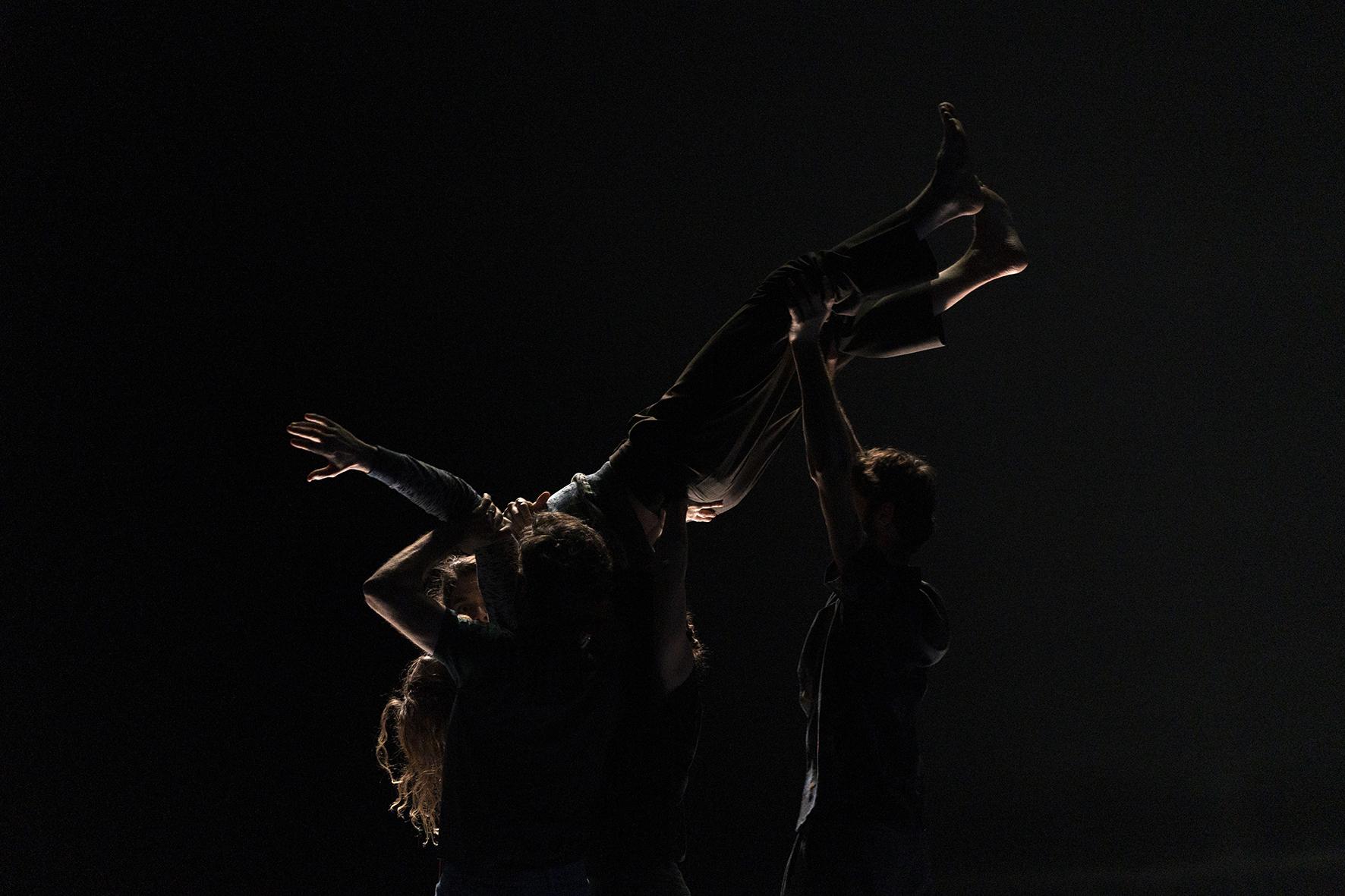 在黑暗的舞台上，舞者們將一位舞者高舉到空中