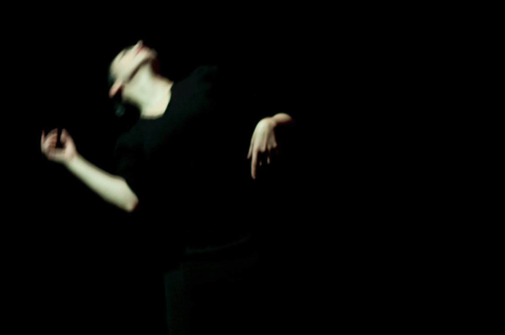 Dancer dressed in black on a black background, head tilted back, blurred in movement