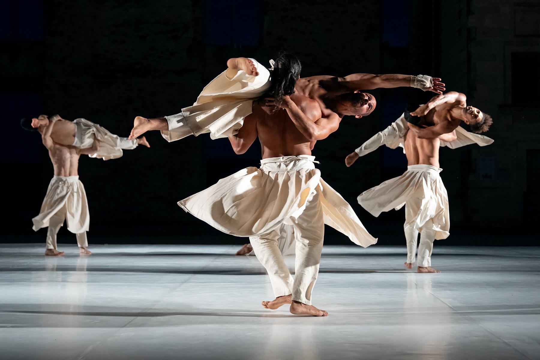 trois hommes en sarouels blancs tournant sur eux-mêmes portent chacun un danseur à l’horizontal