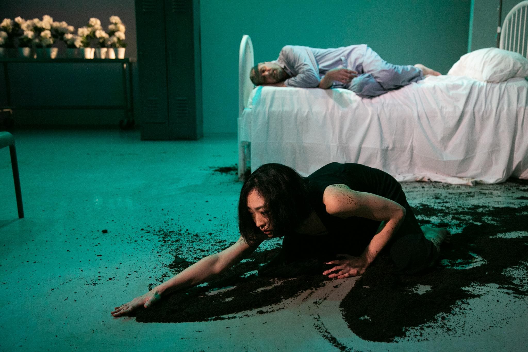 一名男子躺在医院病床上，身旁是一名身穿黑裙的女子，她正在将尘土撒向地板