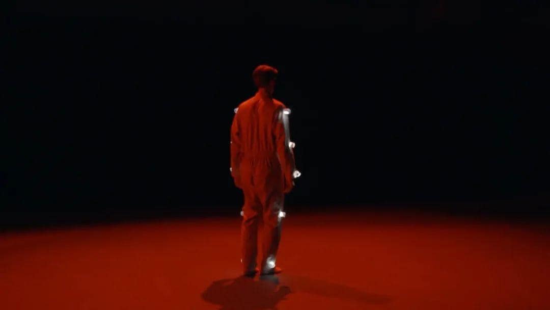 一名身穿带有灯光表演服的男子，站在被红色灯光笼罩着的舞台上，面对舞台后方