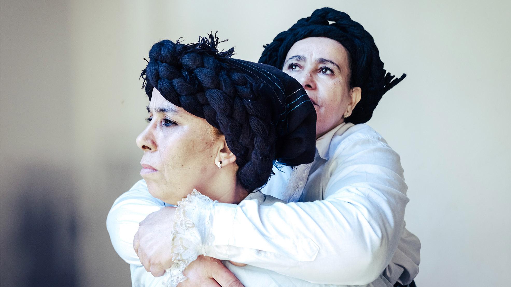 A woman in profile with a black scarf holding her hair and a white shirt, carries on her back another woman dressed identically.