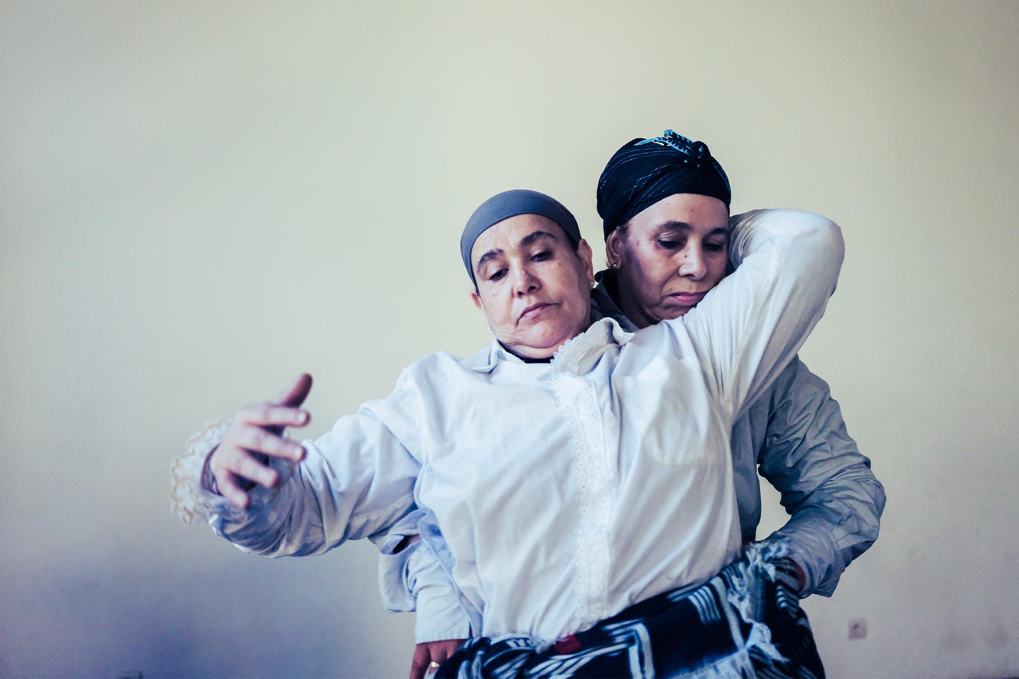 A woman wearing a white shirt, left arm up holding a second woman by her upper back.