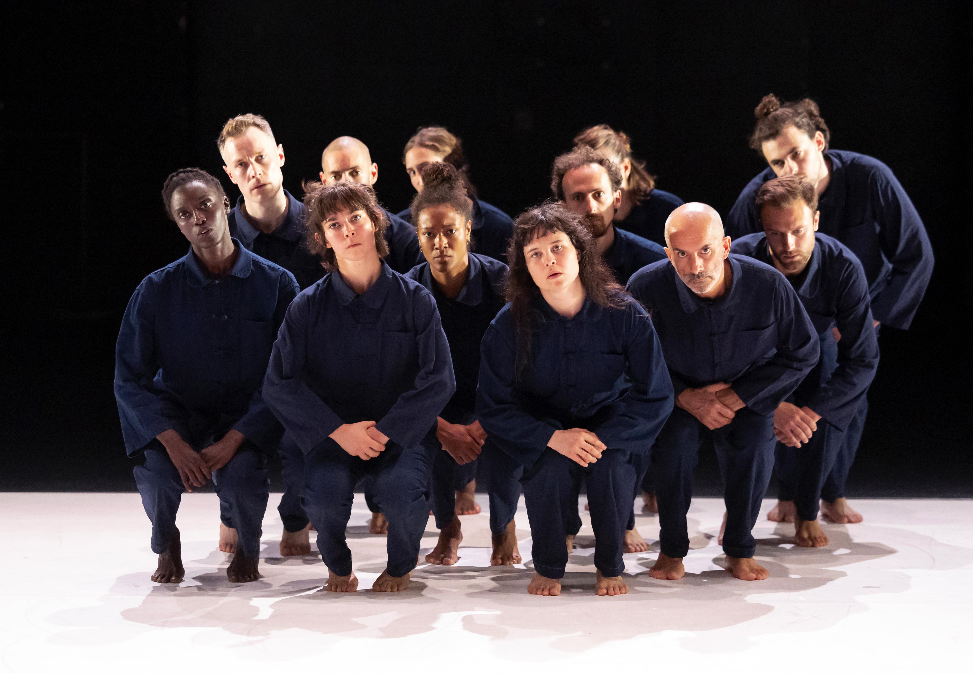 : dancers dressed in overalls, grouped and squatting, hands joined on the knees.