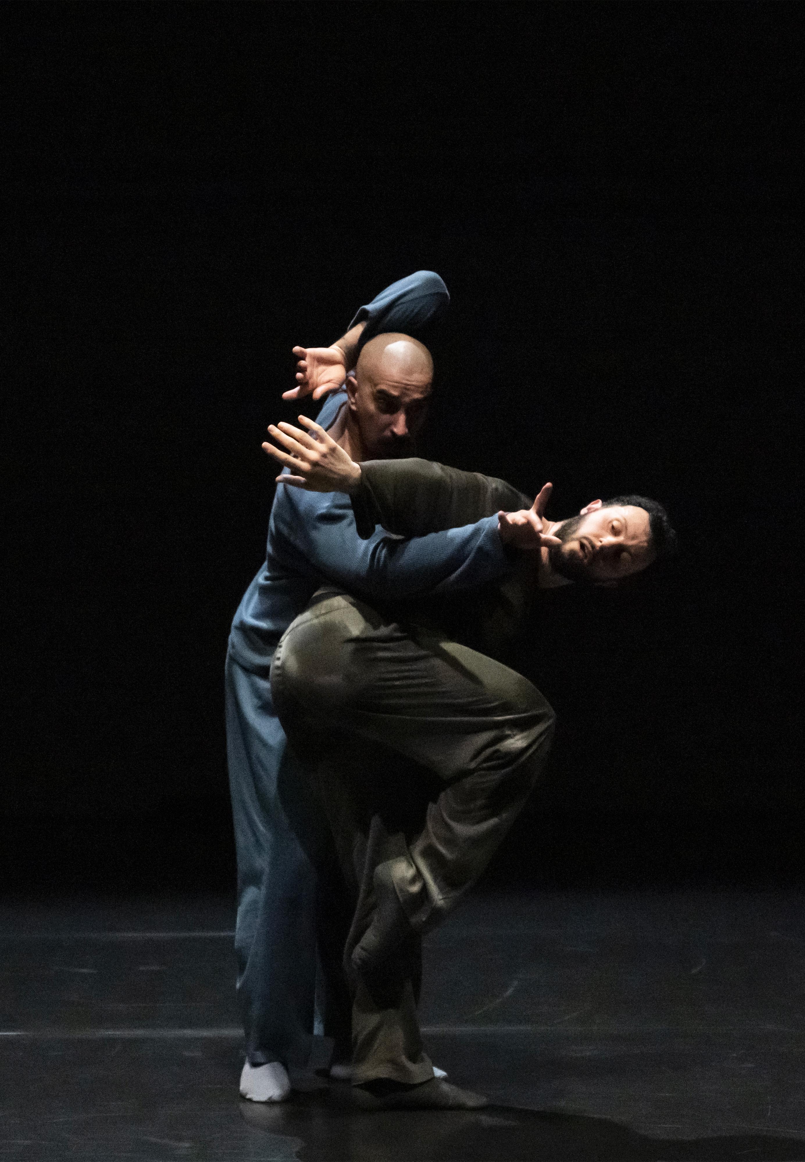 Two dancers side by side in profile, one leaning forward and the other stretching his arm towards the audience