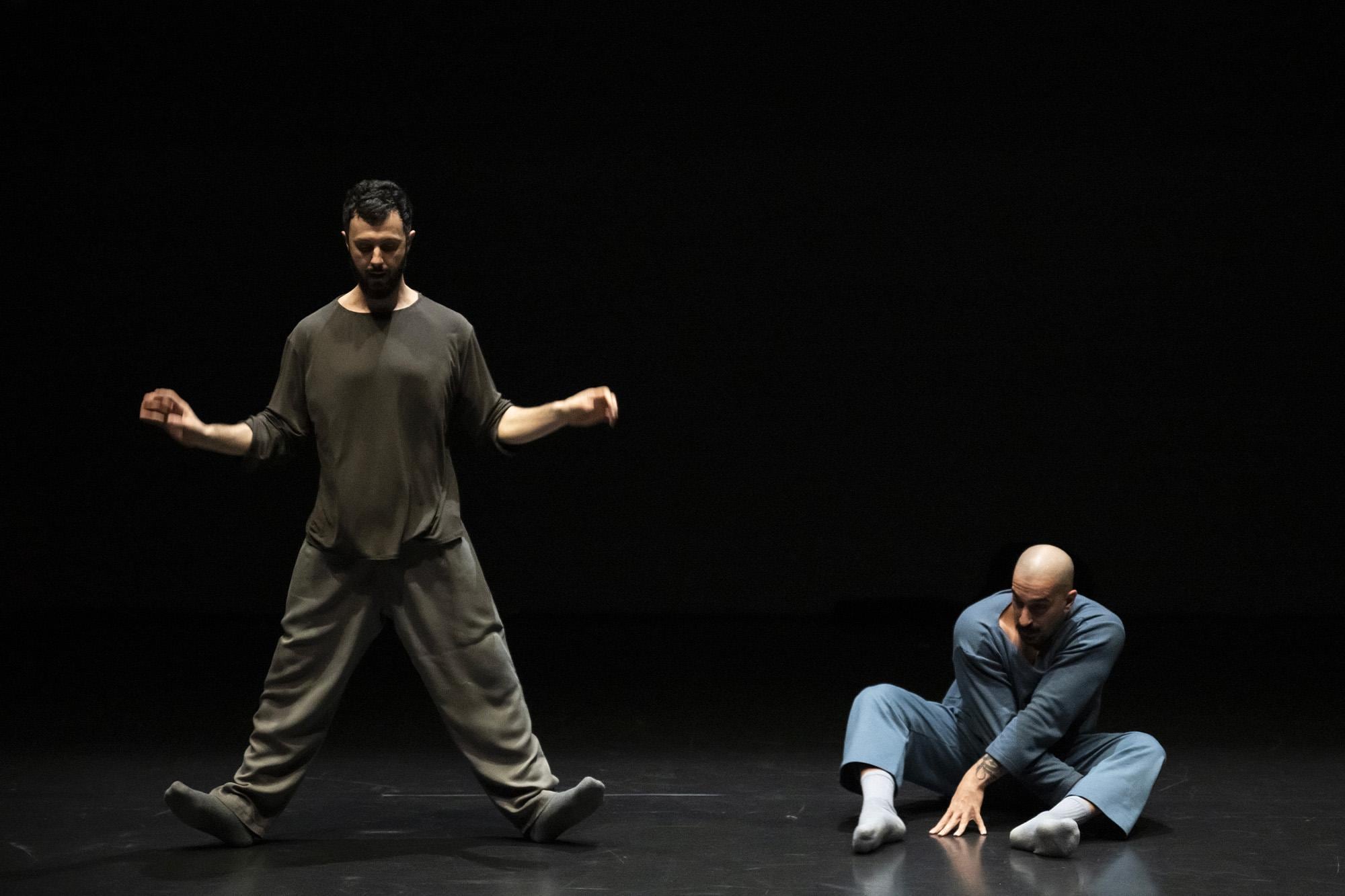 A dancer dressed in khaki, legs spread, leaning on his heels, next to another dancer sitting cross-legged on the floor