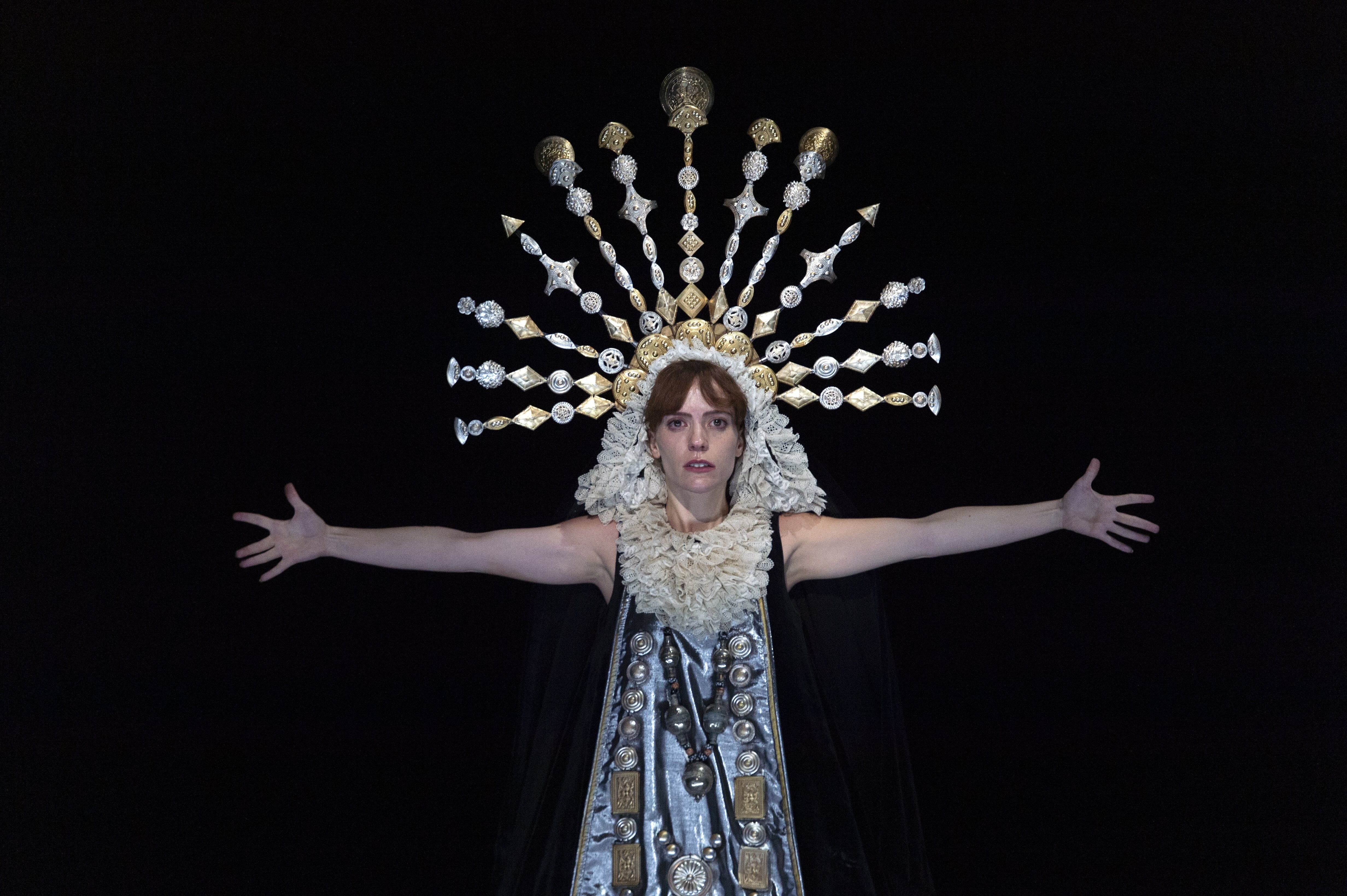 Woman in darkness whose face is bathed in light, wearing a crown in the shape of a halo, with her arms opened.  