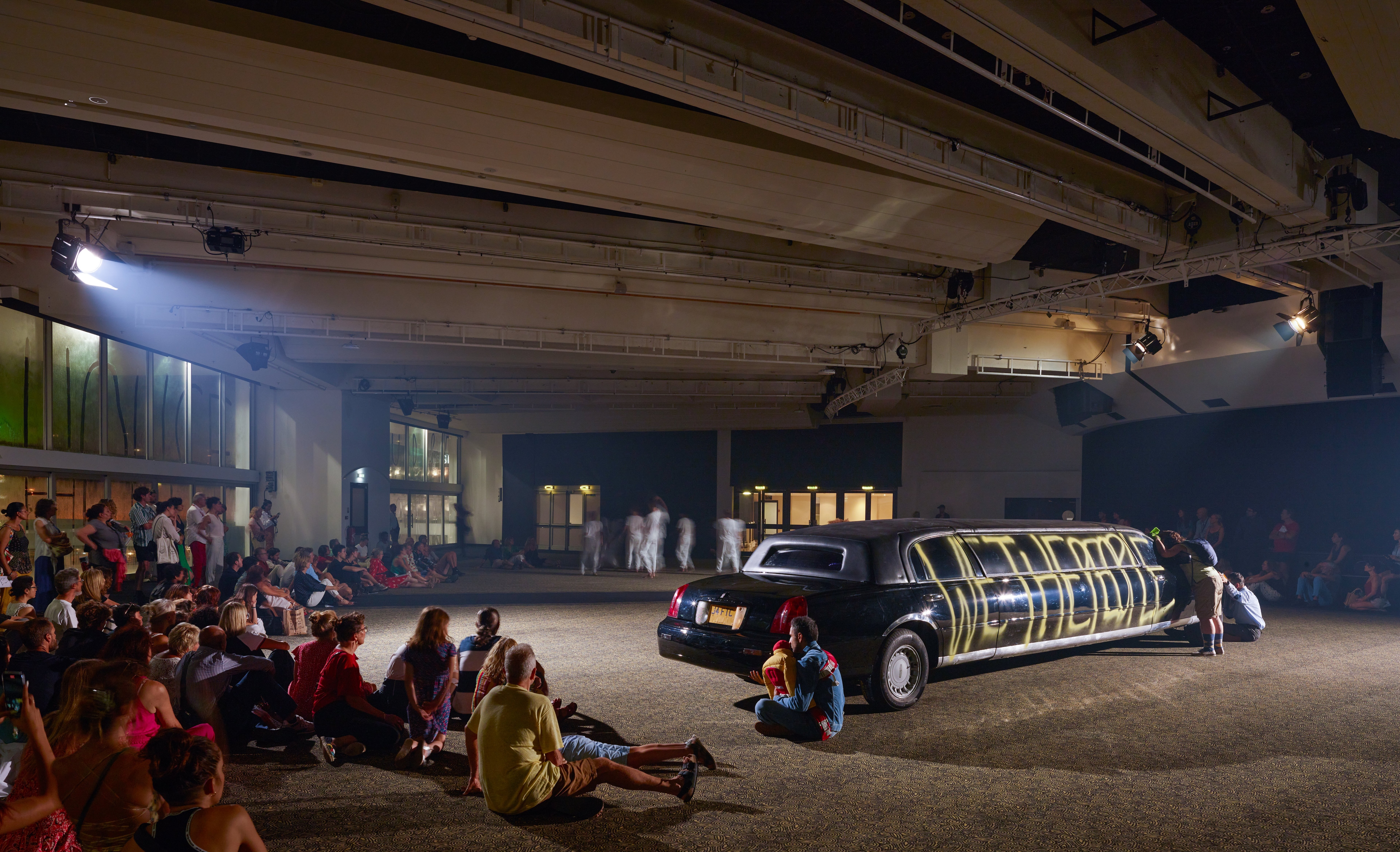 audience sitting on the floor in front of a black limousine