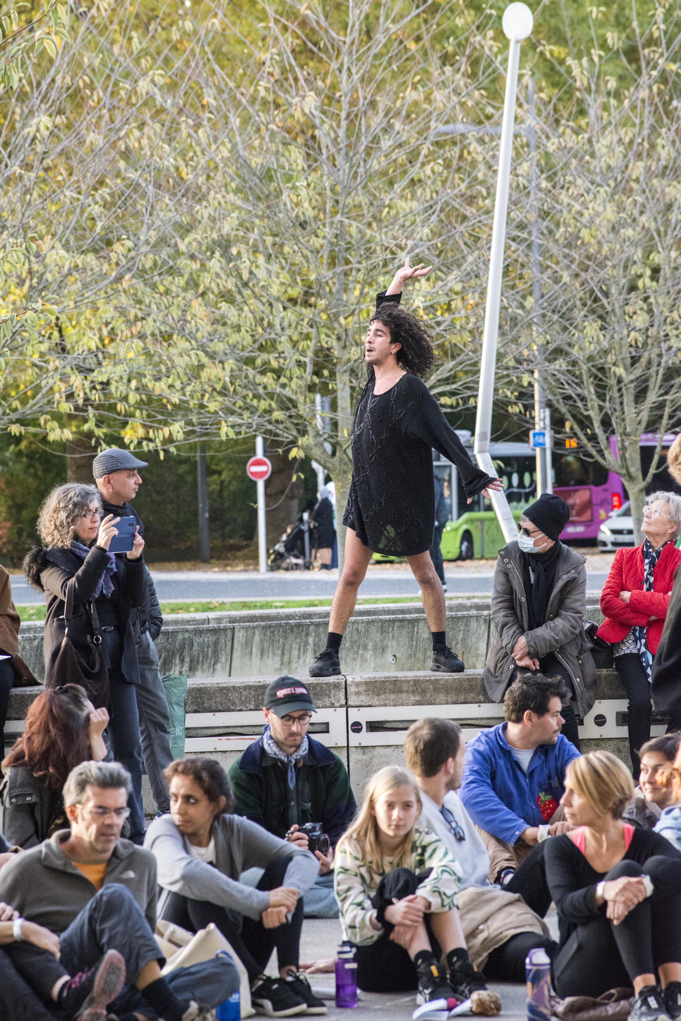 Dancer dressed in a black tunic standing on a concrete wall in the middle of the audience, who are sitting cross-legged