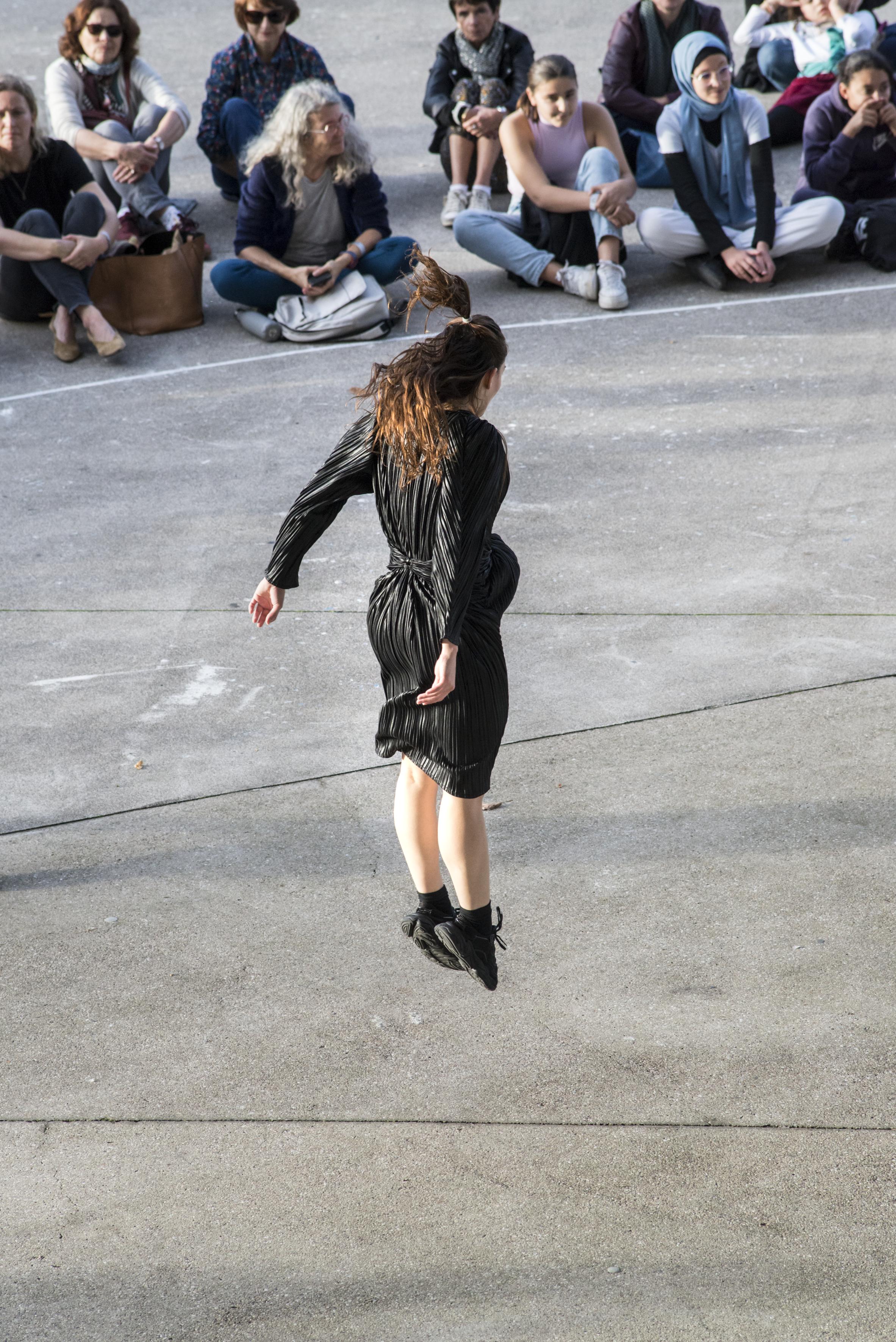 身穿黑色連身裙的舞者在龐畢度中心梅斯分館的廣場上跳躍，面對盤腿而坐的公眾