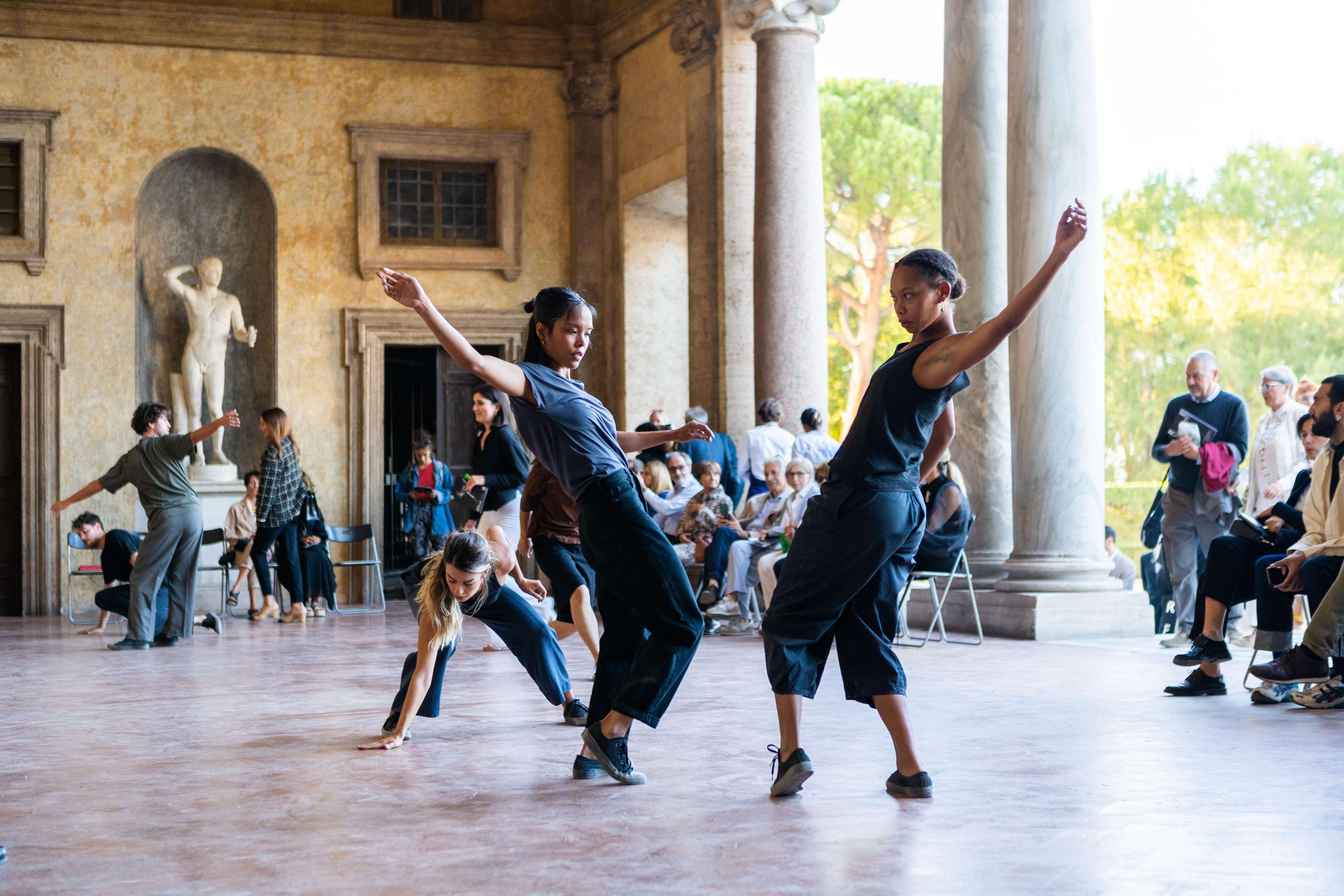 Dancer dressed in black with her right arm in the air and face turned towards the ground, facing another dancer dressed in black with her left arm in the air and her face turned towards the ground – in the Loggia of the Villa Medici