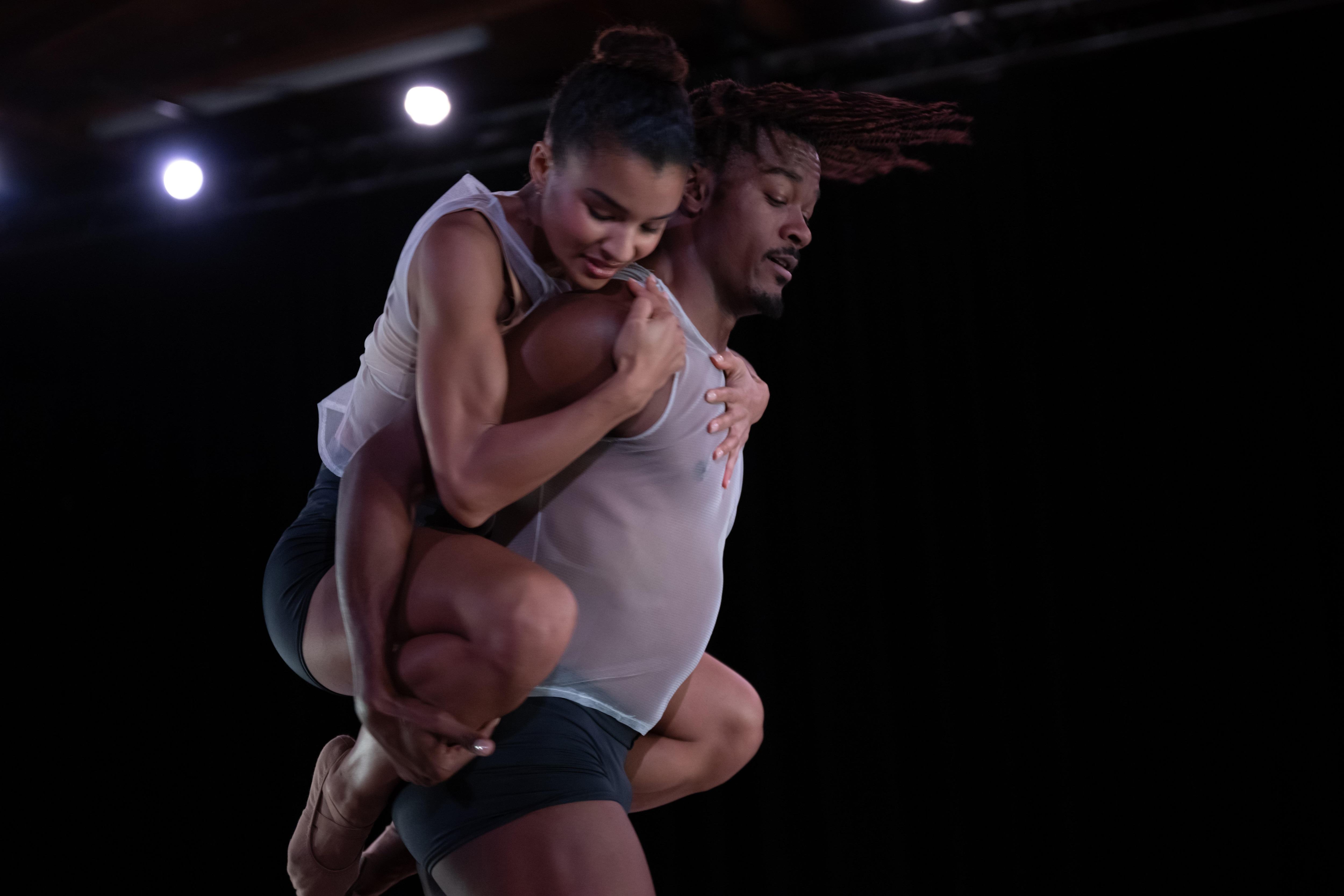 dancer carrying another dancer on his back, both wearing transparent tank tops and black shorts