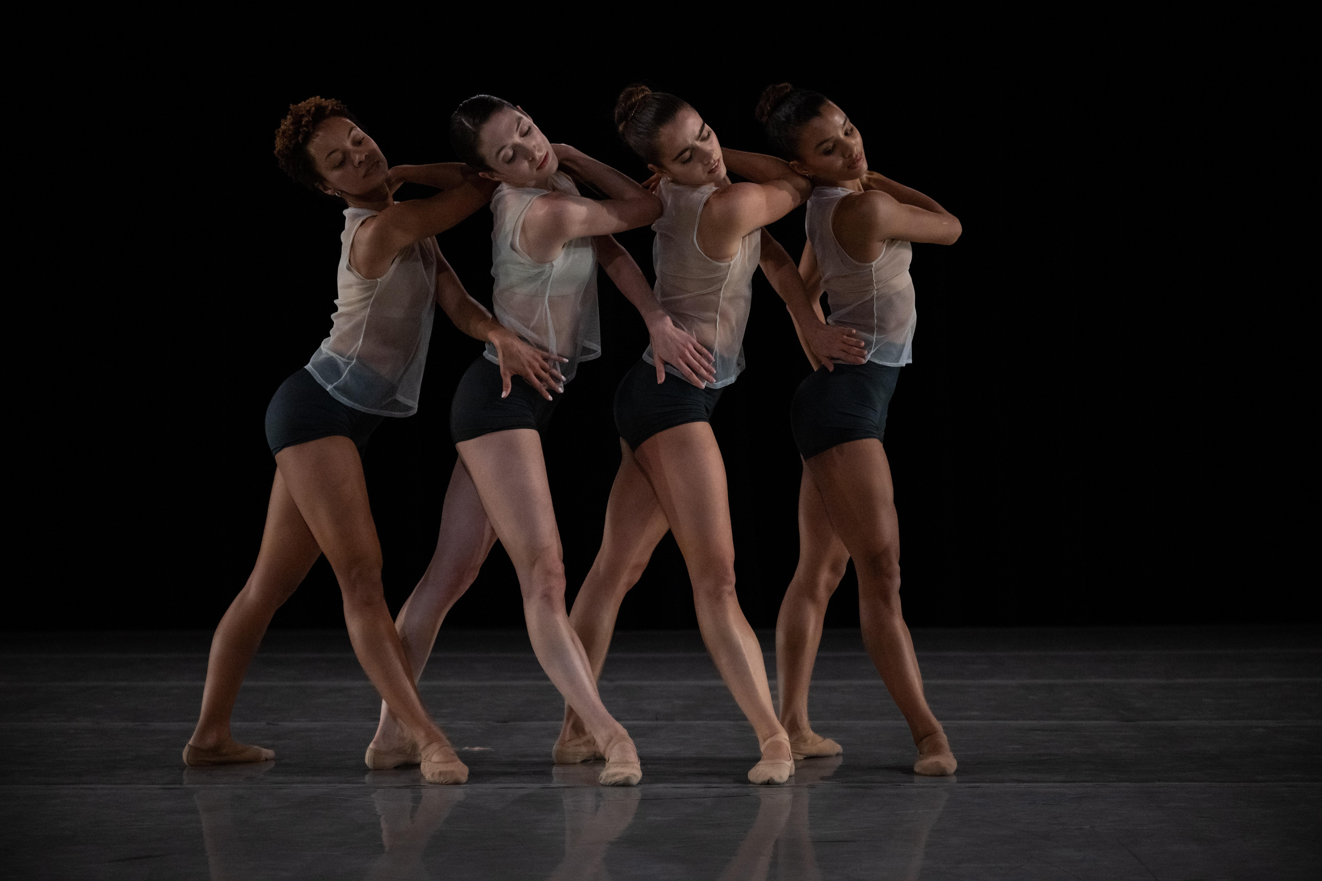 dancers wearing transparent tank tops and black shorts, each behind the other, one hand on their shoulder and the other on the hip of the dancer in front of them.