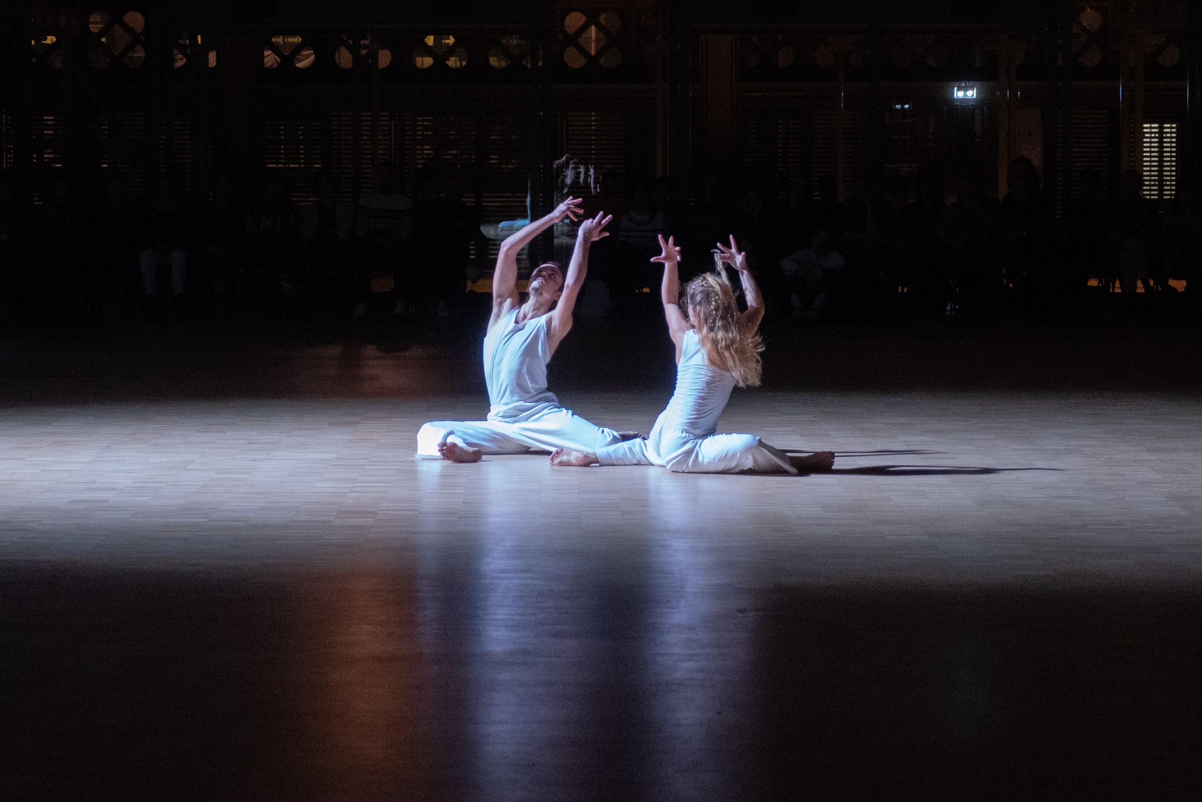 两名舞者面对面坐在地板上，手臂高举