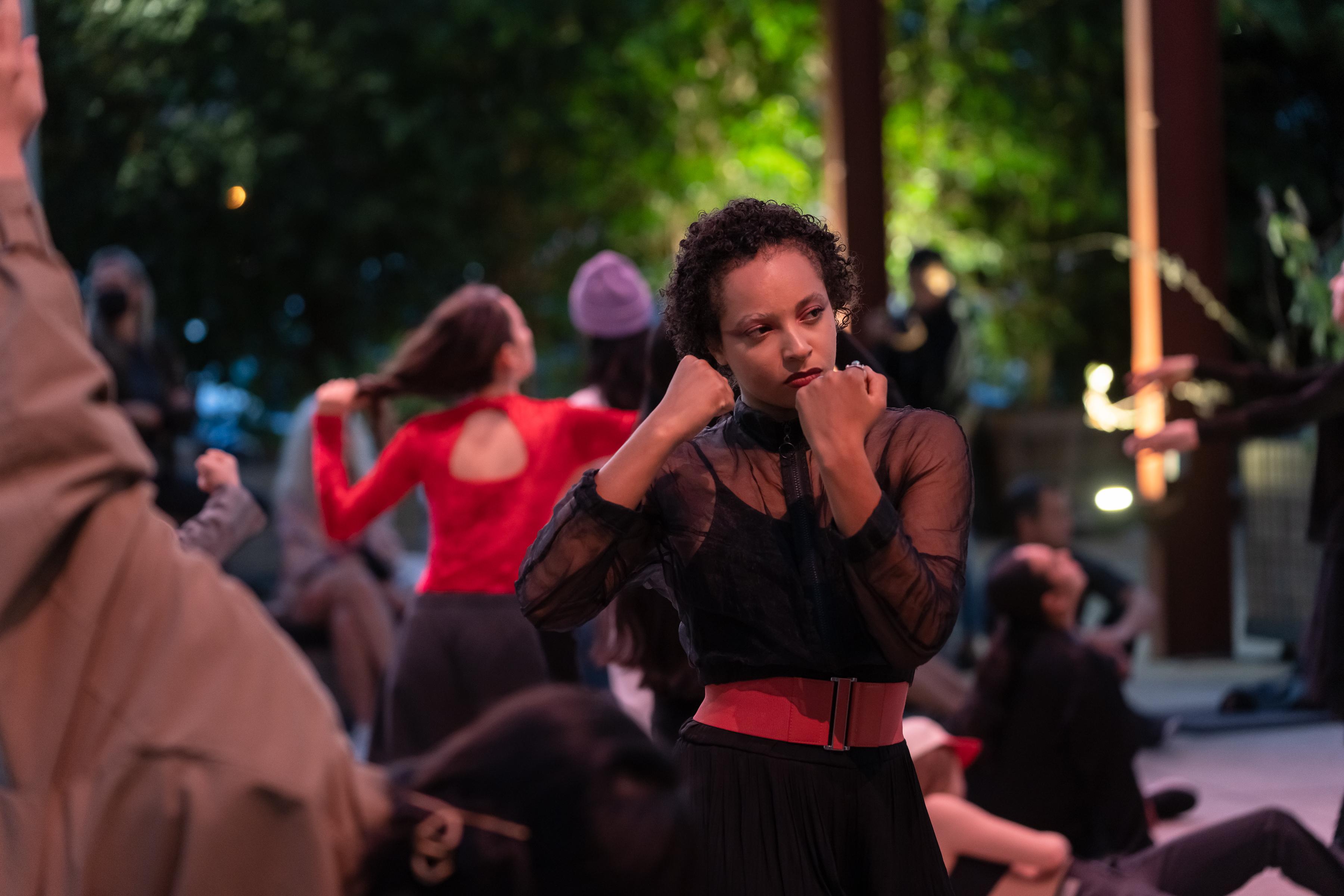 woman wearing a black dress and a red belt around her waist, fists clenched in front of her face