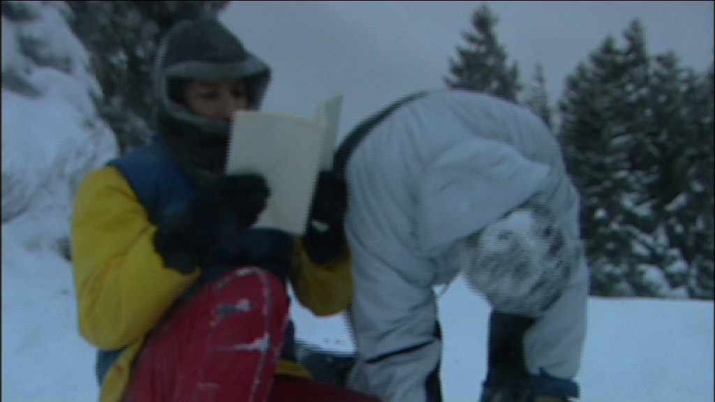 two men dressed warmly, one holding a book, the other with his hands on the ground.